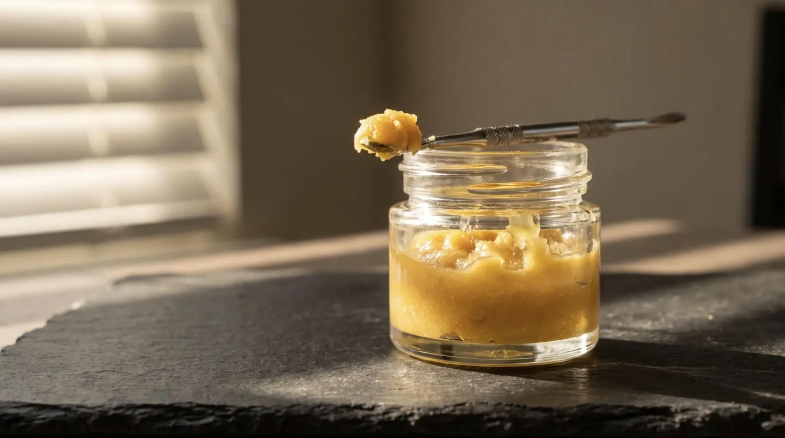 Jar of golden cannabis badder with a dab tool on slate in window light