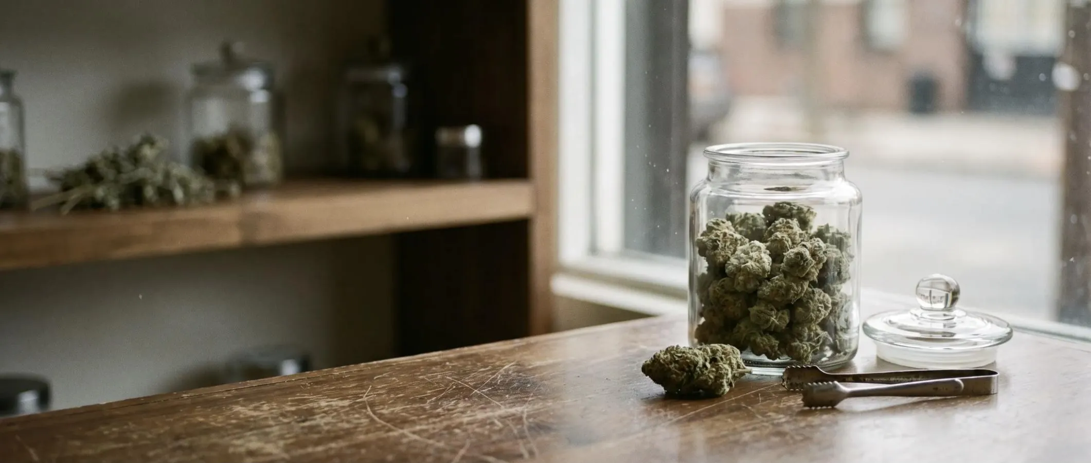 Jar of cannabis flower and steel tongs on a worn wooden shop counter